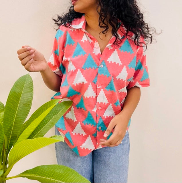 A woman wearing the Peach Puzzle Summer Shirt, featuring a pattern of blue and white triangles. She is smiling and standing near a plant, with the short-sleeved, button-down shirt styled casually with jeans.