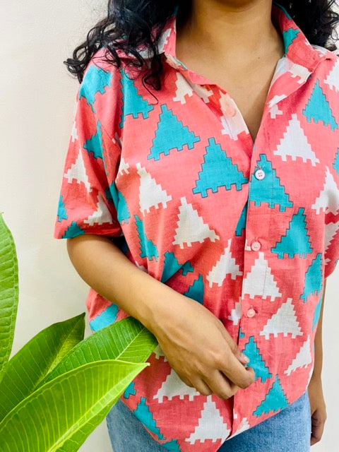 A woman wearing the Peach Puzzle Summer Shirt, featuring a pattern of blue and white triangles. She is smiling and standing near a plant, with the short-sleeved, button-down shirt styled casually with jeans, front close view.