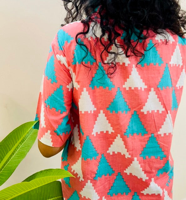 A woman wearing the Peach Puzzle Summer Shirt, featuring a pattern of blue and white triangles. She is smiling and standing near a plant, with the short-sleeved, button-down shirt styled casually with jeans, back close view.