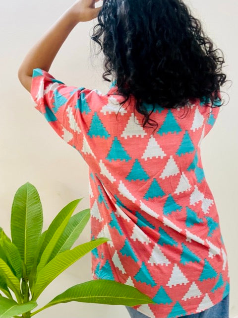 A woman wearing the Peach Puzzle Summer Shirt, featuring a pattern of blue and white triangles. She is smiling and standing near a plant, with the short-sleeved, button-down shirt styled casually with jeans, back view.