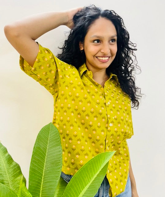 A woman wearing the daisy Summer Shirt, featuring a pattern of daisy. She is smiling and standing near a plant, with the short-sleeved, button-down shirt styled casually with jeans.