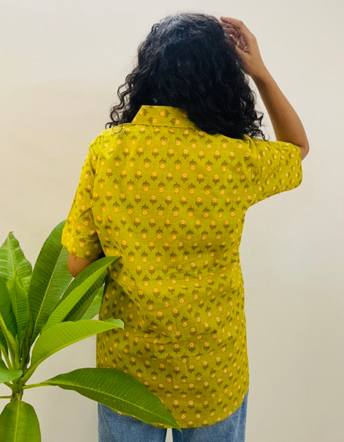 A woman wearing the daisy Summer Shirt, featuring a pattern of daisy. She is smiling and standing near a plant, with the short-sleeved, button-down shirt styled casually with jeans, back view 