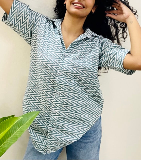 A woman wearing the lazy hazy Summer Shirt, featuring a pattern of lazy hazy. She is smiling and standing near a plant, with the short-sleeved, button-down shirt styled casually with jeans.