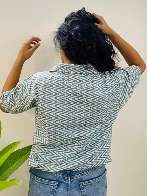 A woman wearing the lazy hazy Summer Shirt, featuring a pattern of lazy hazy. She is smiling and standing near a plant, with the short-sleeved, button-down shirt styled casually with jeans, back view.