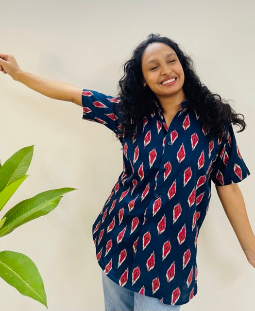 A woman wearing the red ikat Summer Shirt, featuring a pattern of red and white ikat. She is smiling and standing near a plant, with the short-sleeved, button-down shirt styled casually with jeans.