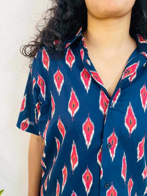 A woman wearing the red ikat Summer Shirt, featuring a pattern of red and white ikat. She is smiling and standing near a plant, with the short-sleeved, button-down shirt styled casually with jeans, close up