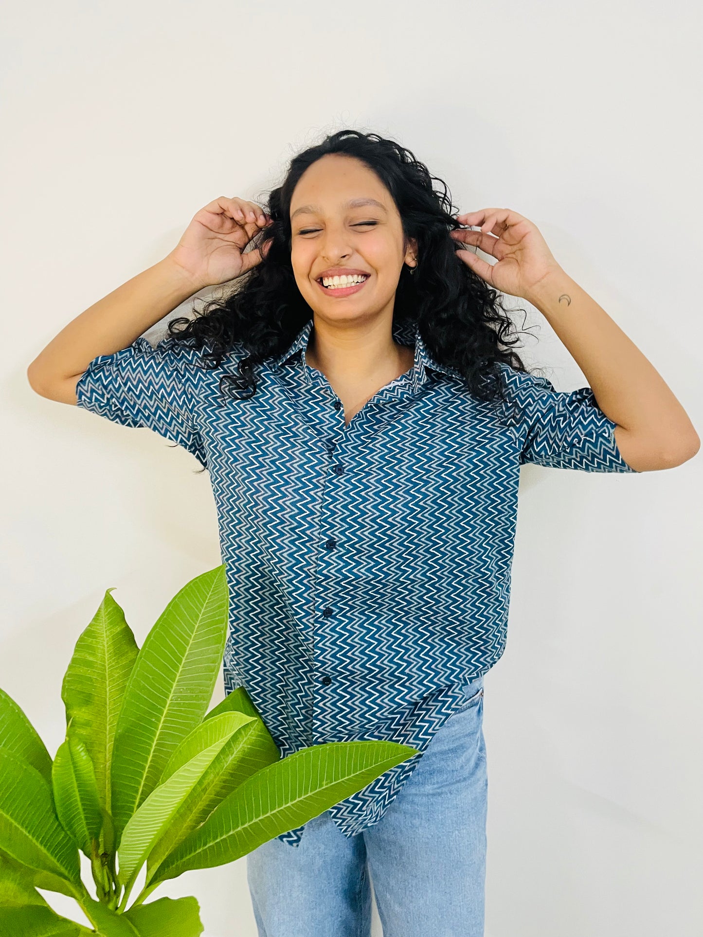 A woman wearing the lazy hazy Summer Shirt, featuring a pattern of lazy hazy. She is smiling and standing near a plant, with the short-sleeved, button-down shirt styled casually with jeans.