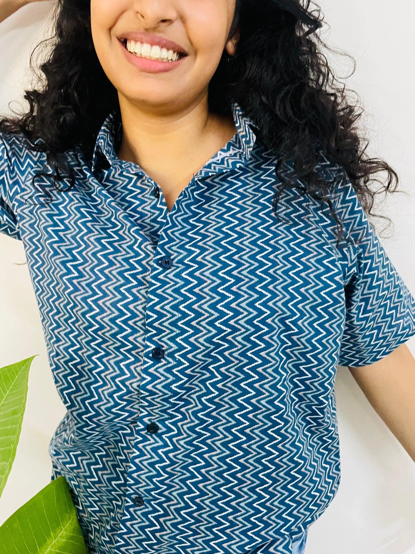 A woman wearing the lazy hazy Summer Shirt, featuring a pattern of lazy hazy. She is smiling and standing near a plant, with the short-sleeved, button-down shirt styled casually with jeans.