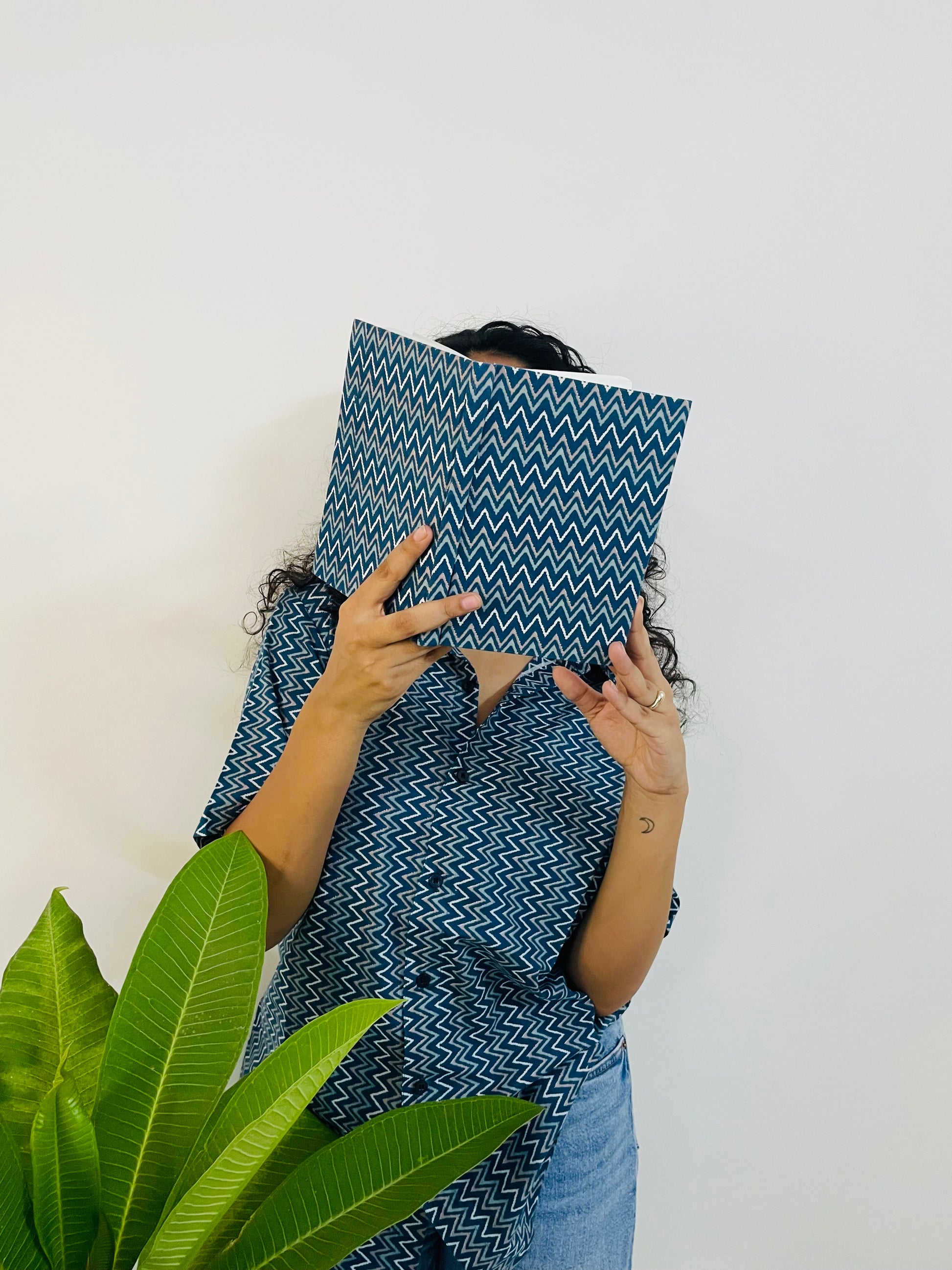 A woman wearing the lazy hazy Summer Shirt, featuring a pattern of lazy hazy. She is smiling and standing near a plant, with the short-sleeved, button-down shirt styled casually with jeans.