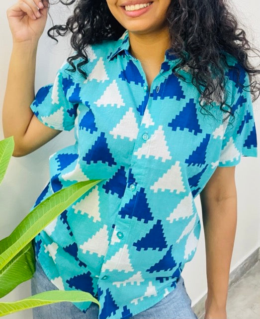 A woman wearing the Blue Puzzle Summer Shirt, featuring a pattern of blue and white triangles. She is smiling and standing near a plant, with the short-sleeved, button-down shirt styled casually with jeans.