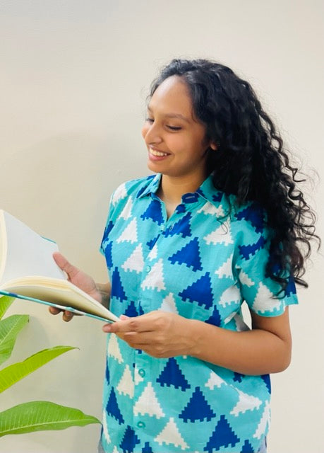 A woman wearing the Blue Puzzle Summer Shirt, featuring a pattern of blue and white triangles. She is smiling and standing near a plant, with the short-sleeved, button-down shirt styled casually with jeans, holding our journal.