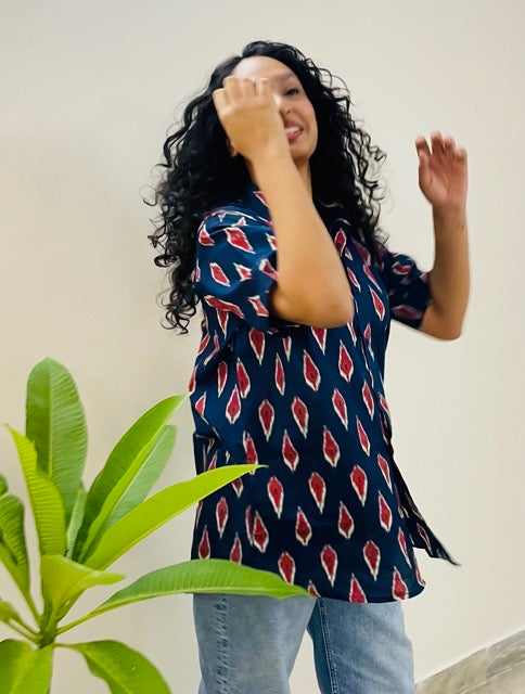 A woman wearing the red ikat Summer Shirt, featuring a pattern of red and white ikat. She is smiling and standing near a plant, with the short-sleeved, button-down shirt styled casually with jeans.