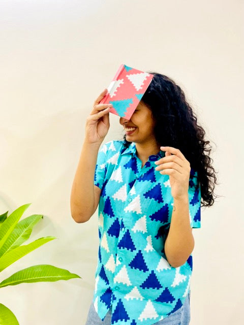 A woman wearing the Blue Puzzle Summer Shirt, featuring a pattern of blue and white triangles. She is smiling and standing near a plant, with the short-sleeved, button-down shirt styled casually with jeans holding peach handmade journal on the head.