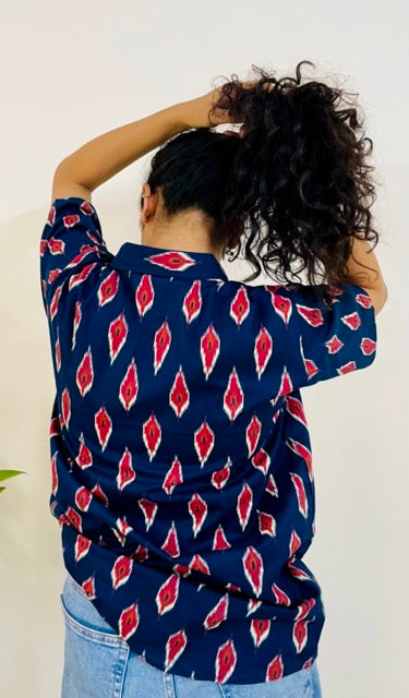 A woman wearing the red ikat Summer Shirt, featuring a pattern of red and white ikat. She is smiling and standing near a plant, with the short-sleeved, button-down shirt styled casually with jeans, back view.