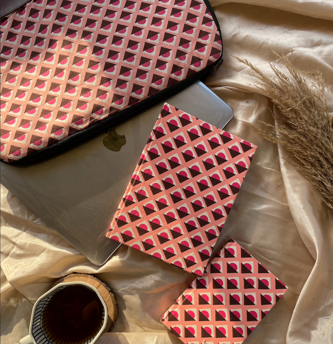 Three products displayed against a light golden cloth background. From left to right: a laptop bag, a 6x8 handmade journal, and a 6x4 handmade journal, all featuring a Geoplay design pattern. A MacBook and a cup of coffee are also visible in the background